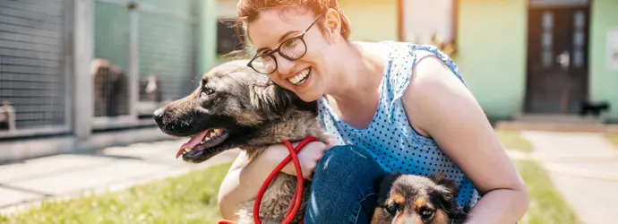 A young woman crouches, hugs her dog in a sunny garden.