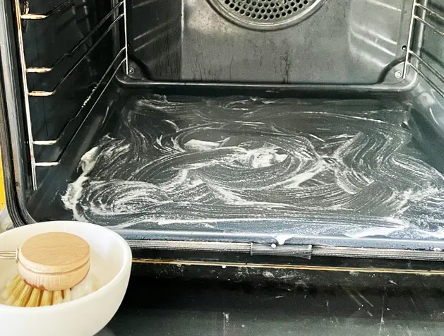 Young woman in apron cleaning the oven with spray and cloth