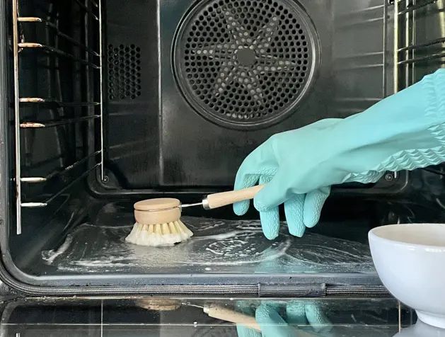 Young woman in apron cleaning the oven with spray and cloth