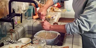 Woman with her sleeves rolled up washing dirty dishes in a full kitchen sink.
