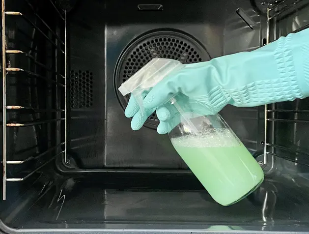 Young woman in apron cleaning the oven with spray and cloth