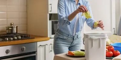 A woman in a blue shirt peels vegetables into a compost bin.