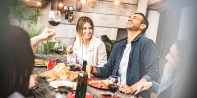 A group of smiling friends enjoying wine and food around a table at a garden party.