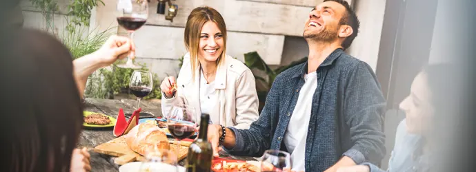 A group of smiling friends enjoying wine and food around a table at a garden party.