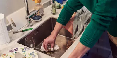 Man in a green jumper cleaning the kitchen sink with some tissue paper.