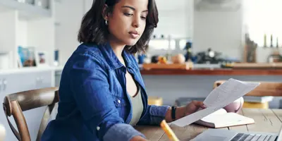 A woman focuses on paperwork at a laptop in a sunny room.