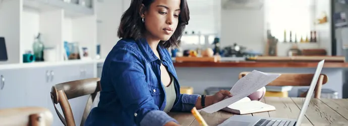 A woman focuses on paperwork at a laptop in a sunny room.