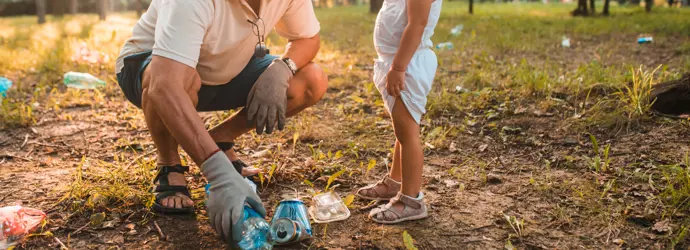 Grandpa teaches toddler to collect litter in a sunlit park.