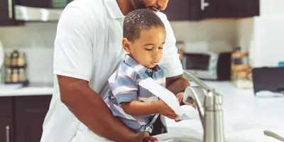 A dad washes hands with his toddler at a kitchen sink.