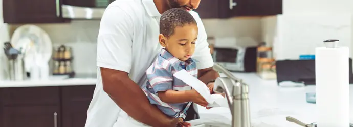 A dad washes hands with his toddler at a kitchen sink.