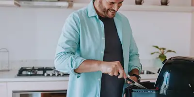 A smiling man stirs food in an airfryer, in a bright kitchen