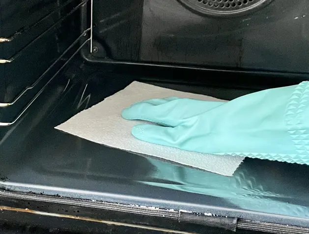 Young woman in apron cleaning the oven with spray and cloth