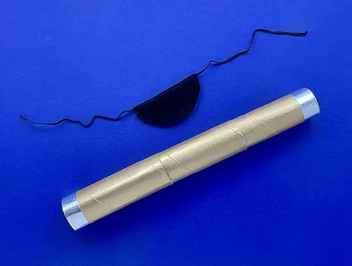 The gold DIY telescope is laid on a blue background below a black DIY fabric and elastic eye patch.