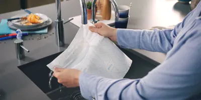 Man in a blue shirt holds a sheet of Plenty kitchen paper under a stainless still kitchen sink tap