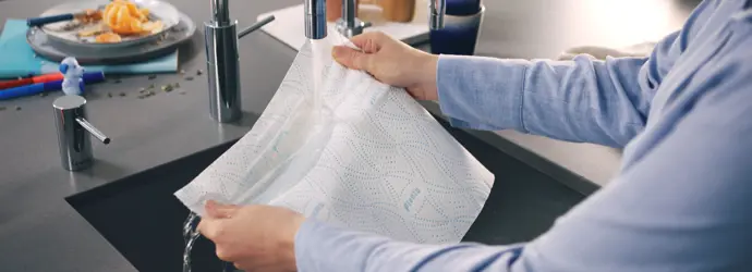 Man in a blue shirt holds a sheet of Plenty kitchen paper under a stainless still kitchen sink tap